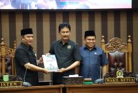 Plt Asisten Pemerintahan dan Kesra, Wisnu Wardhana (kiri), menyerahkan dokumen Raperda Pembentukan Desa Baru kepada Ketua Bapemperda DPRD Tanah Bumbu, H. Harmanudin (tengah), disaksikan oleh Wakil Ketua I DPRD Tanah Bumbu, H. Hasanuddin (kanan), dalam Rapat Paripurna DPRD Tanah Bumbu, Senin (6/10/2025).