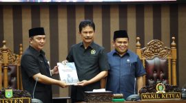 Plt Asisten Pemerintahan dan Kesra, Wisnu Wardhana (kiri), menyerahkan dokumen Raperda Pembentukan Desa Baru kepada Ketua Bapemperda DPRD Tanah Bumbu, H. Harmanudin (tengah), disaksikan oleh Wakil Ketua I DPRD Tanah Bumbu, H. Hasanuddin (kanan), dalam Rapat Paripurna DPRD Tanah Bumbu, Senin (6/10/2025).