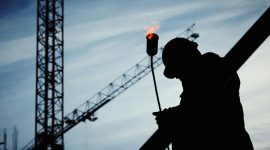 Ilustrasi pekerja konstruksi tengah menggunakan alat pembakar di lokasi proyek dengan latar belakang crane, menggambarkan risiko kerja tinggi yang dihadapi pekerja di sektor konstruksi.