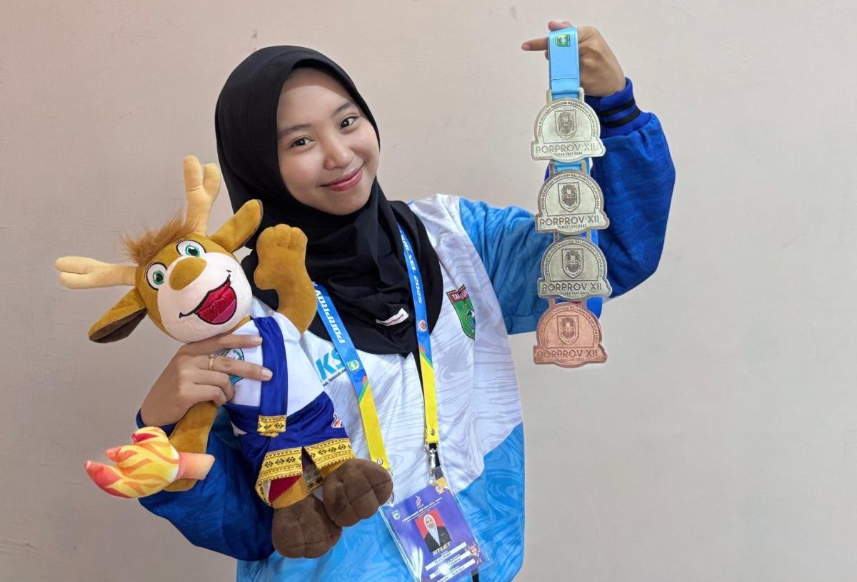 Muslimah mengenakan jaket kontingen olahraga tampak tersenyum sambil memegang beberapa medali PORPROV XII dan boneka maskot kegiatan, menunjukkan kebanggaan atas prestasi yang diraihnya, Jumat (28/11/2025)..